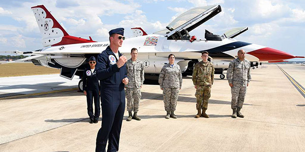 Air Force personnel in flight suits and uniforms stand on an airfield apron with a red, white, and blue Thunderbird F-16 jet behind them.